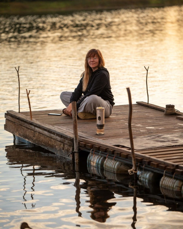 Woman in a dark jacket sitting at the end of a wooden dock on a lake at golden hour, a Brulé espresso maker standing on the planks beside her