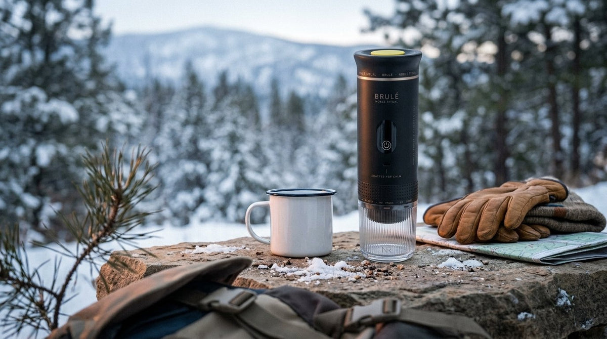 Black Brulé espresso maker on a snowy stone ledge beside an enamel mug, leather gloves and a trail map