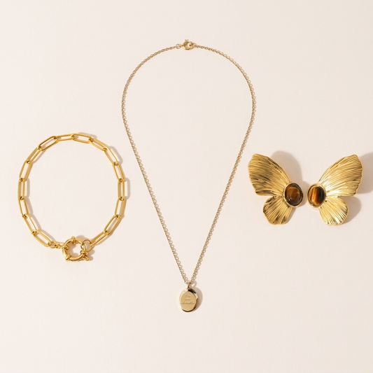 A gold-tone necklace with a small oval pendant, a pair of gold-tone butterfly earrings with brown stones and a gold-tone paperclip-link bracelet on a warm pearl background